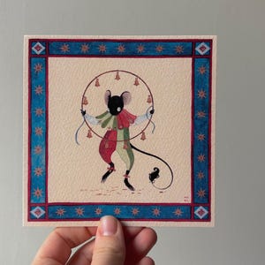 Puede incluir: Ilustración en acuarela de un ratón con un traje de arlequín, sosteniendo un círculo de campanas. La obra de arte está enmarcada con un borde azul y rojo con detalles de estrellas. El ratón tiene una larga cola y está sobre un fondo crema.