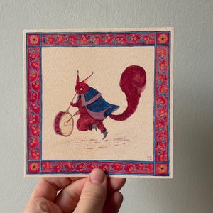 May include: Square watercolor painting of a red creature playing a drum, framed by a red and blue floral border. The creature wears a blue cape and has a large, fluffy tail. The drum is beige.
