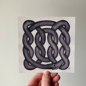 May include: A watercolour painting of a stylised snake in a knot design. The snake is dark grey with a textured look, and it has a smiling face with white teeth and black eyes. The artwork is held by a hand.