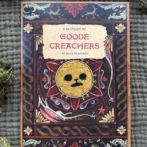 May include: A book titled "A Bestiary of Goode Creachers" by Hana Berggren. The cover features a gold sun-like design with a face, surrounded by a decorative border in shades of brown, red, and gold. The book is on a green surface.