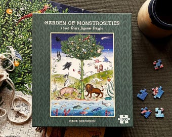 May include: A 1000-piece jigsaw puzzle titled "Garden of Monstrosities" with a whimsical illustration of a tree, animals, and fish. The box is green with a patterned border. Scattered puzzle pieces and a mug are also visible.