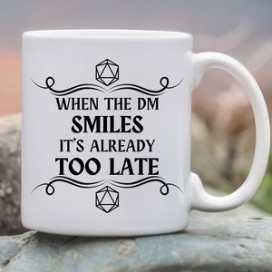 May include: White ceramic mug with a black design that says "When the DM smiles it's already too late". The design also includes a stylized 20-sided die.