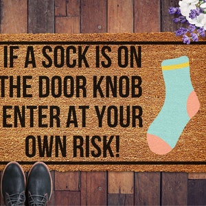 May include: A brown doormat with the text "If a sock is on the door knob enter at your own risk!" in black lettering. A blue sock with a yellow stripe and pink heel is on the right side of the doormat.
