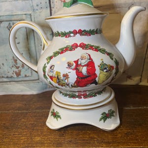 Könnte beinhalten: Eine weiße Keramik-Teekanne mit Weihnachtsmotiv. Die Teekanne zeigt eine Illustration des Weihnachtsmanns, Stechpalmen- und Schleifenakzente und goldfarbene Verzierungen. Der Deckel ist mit Stechpalmen und Beeren verziert. Die Teekanne steht auf einem passenden Sockel.