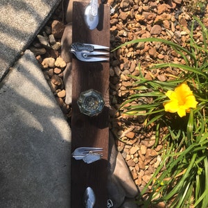 Puede incluir: Un llavero rústico de pared de madera con cubiertos reciclados. El soporte incluye una cuchara, un tenedor y otros utensilios, cada uno sirviendo como gancho. La madera es de color marrón oscuro y los cubiertos son plateados. El llavero se exhibe al aire libre.