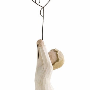 Könnte beinhalten: Eine geschnitzte Holzfigur einer Person, die einen Drahtballon hält. Der Ballon ist schwarz und trägt das Wort "Congrats" in Schreibschrift. Die Figur ist hellbeige und hat eine strukturierte Oberfläche.