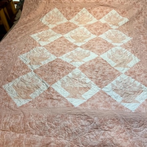 May include: A quilt featuring a repeating diamond pattern in shades of dusty rose and white. The fabric displays a subtle floral print, and the surface is finished with intricate, swirling machine-stitched quilting patterns throughout the entire piece.