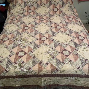 Op de afbeelding: Een beige quilt met patroon en een geometrisch ontwerp. De quilt heeft een patchwork van driehoeken, vierkanten en bloempatronen in tinten beige, roze, bruin en grijs. De randen zijn afgewerkt met een donkerbruine rand.