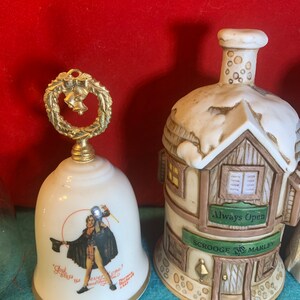 May include: Two porcelain Christmas bells. One bell is white with a gold trim and features a depiction of Ebenezer Scrooge from A Christmas Carol. The other bell is shaped like a building with a sign that reads "Scrooge & Marley" and "Always Open".
