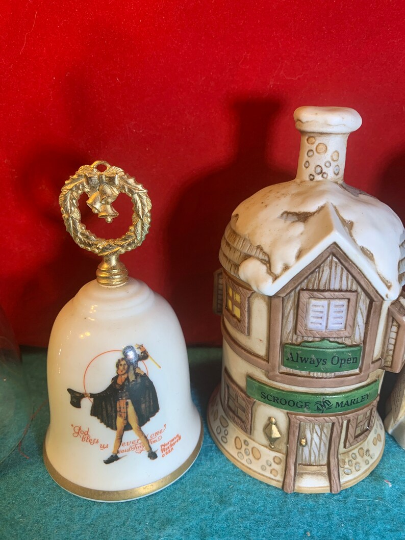 May include: Two porcelain Christmas bells. One bell is white with a gold trim and features a depiction of Ebenezer Scrooge from A Christmas Carol. The other bell is shaped like a building with a sign that reads "Scrooge & Marley" and "Always Open".