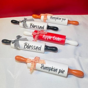 May include: Four white rolling pins with black handles and orange handles. The rolling pins are decorated with black and white checkered ribbon and orange and white checkered ribbon. The rolling pins are labeled "Pumpkin Pie", "Blessed", "Apple Cider", and "Blessed".