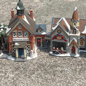Può includere: Una collezione di edifici in ceramica in miniatura, tra cui negozi e chiese, con dettagli architettonici. Gli edifici hanno schemi di colore rosso, marrone e bianco, con tetti innevati e accenti decorativi. Gli edifici sono disposti in fila.