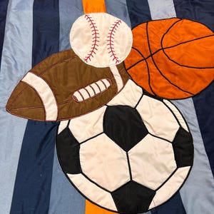 Könnte beinhalten: Eine Bettbezug im Sport-Design mit aufgenähten Bildern eines Baseballs, Footballs, Basketballs und Fußballs. Der Hintergrund hat vertikale Streifen in Blautönen und einen orangefarbenen Akzentstreifen.