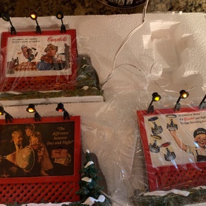 May include: Three miniature red and white billboard signs with light bulbs on top. The signs feature vintage advertisements for Campbell's Soup, a globe, and a bottle of sparkling water. The signs are on a white surface with a snowy background.