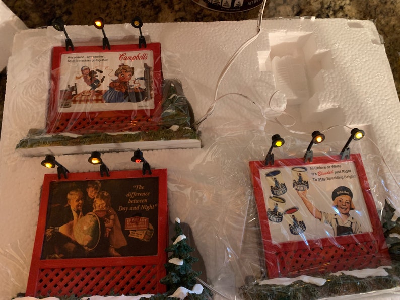 May include: Three miniature red and white billboard signs with light bulbs on top. The signs feature vintage advertisements for Campbell's Soup, a globe, and a bottle of sparkling water. The signs are on a white surface with a snowy background.