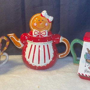 Puede incluir: Tres teteras de cerámica con motivos navideños. Una es un muñeco de nieve con sombrero y bufanda azules, otra es un hombre de jengibre, y la tercera tiene una tapa roja y un asa verde con el texto "Pass the Tea".
