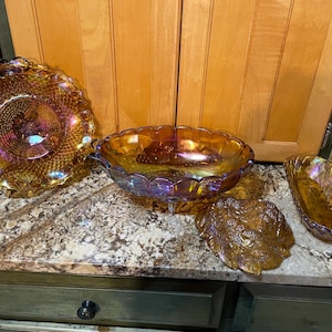 May include: An assortment of iridescent amber glass serving dishes. The collection includes a large round plate with a decorative edge, an oval bowl with handles, a leaf-shaped dish, and a small oval dish. The glass has a shimmering, colorful finish.