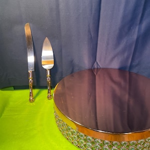 May include: A cake stand with a mirrored top and a gold-colored rim adorned with clear, round crystals. A matching cake knife and server with gold-colored handles are also present. The items are on a green surface.