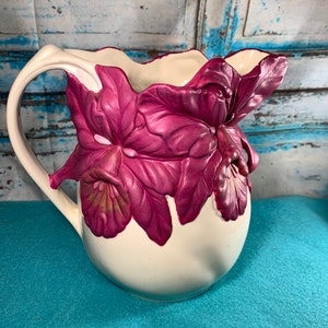 May include: A white ceramic pitcher with a pink floral design. The pitcher has a wide mouth and a handle. The flowers are three-dimensional and have a realistic appearance.