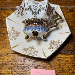 Może przedstawiać: Zestaw filiżanki i spodka do herbaty w stylu vintage. Biała porcelanowa filiżanka ma złote wykończenia, niebiesko-złoty ozdobny pasek i złote akcenty. Spodek jest biały ze złotymi wykończeniami i wzorem koniczyny. Zestaw stoi na drewnianej powierzchni.