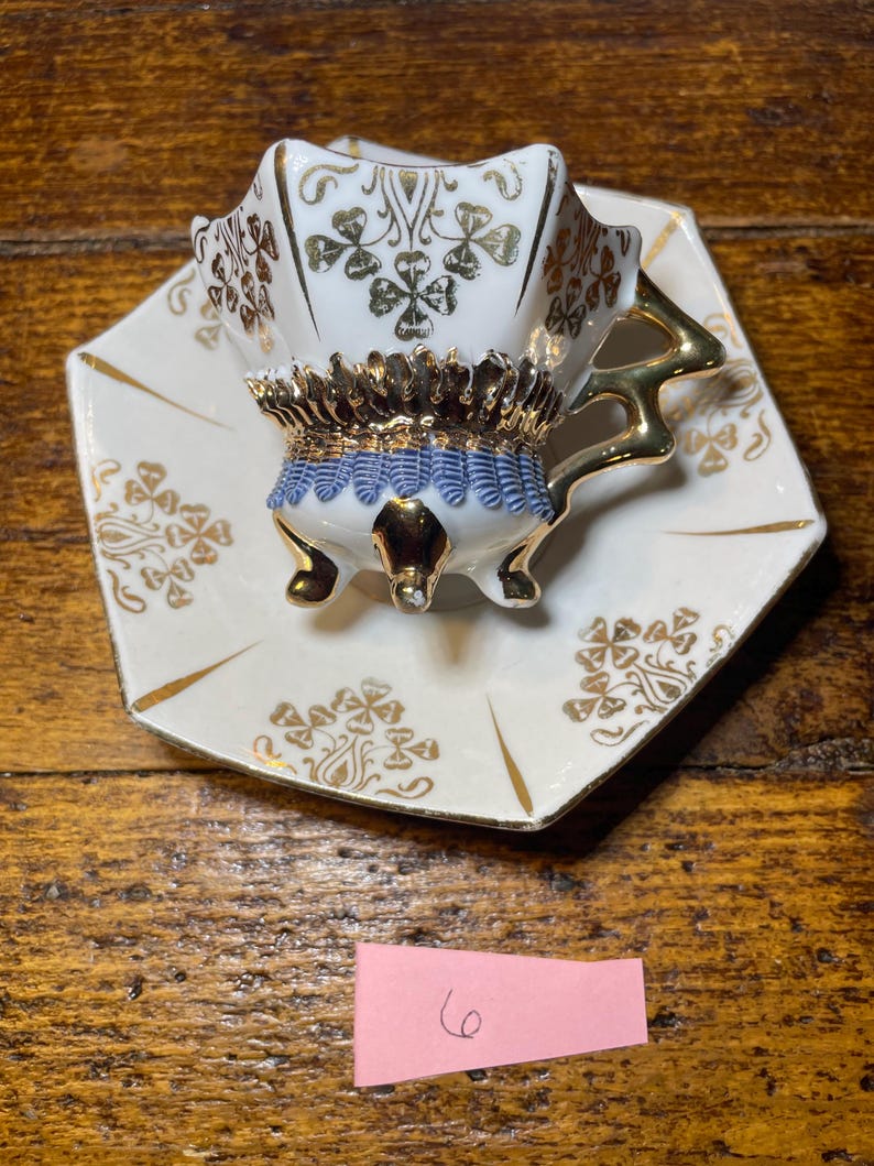 Może przedstawiać: Zestaw filiżanki i spodka do herbaty w stylu vintage. Biała porcelanowa filiżanka ma złote wykończenia, niebiesko-złoty ozdobny pasek i złote akcenty. Spodek jest biały ze złotymi wykończeniami i wzorem koniczyny. Zestaw stoi na drewnianej powierzchni.