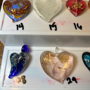 May include: A collection of heart-shaped glass pendants in various colors and designs. The pendants include clear, red, blue, and gold accents. Each pendant has a loop for hanging and is displayed on a white surface with numbers.