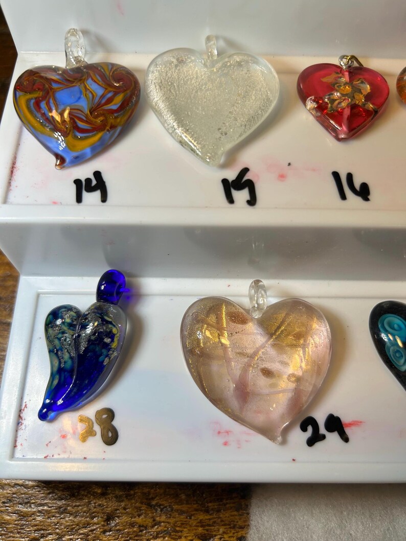 May include: A collection of heart-shaped glass pendants in various colors and designs. The pendants include clear, red, blue, and gold accents. Each pendant has a loop for hanging and is displayed on a white surface with numbers.