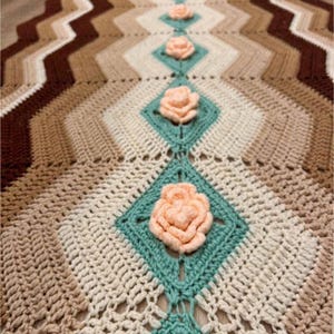 Pode incluir: Caminho de mesa de crochê com padrão chevron em tons de castanho, bege e creme. Apresenta detalhes de diamantes turquesa e aplicações de rosas pêssego. Um têxtil decorativo para casa, com cerca de 150 cm de comprimento.