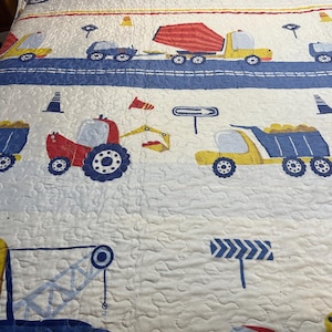 May include: A white quilted bedspread featuring a repeating pattern of construction vehicles including yellow dump trucks, red tractors, and blue cranes. The design includes blue road stripes, orange traffic cones, and directional road signs.
