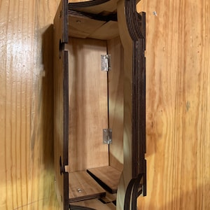 Puede incluir: Un mueble de madera para colgar en la pared con un acabado marrón claro y bordes marrones oscuros. El mueble tiene forma rectangular con detalles curvos decorativos y una pequeña balda en el interior. Está fijado a una pared de madera con vetas visibles.