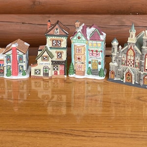 Puede incluir: Una colección de 13 casas de pueblo de Navidad en miniatura hechas de cerámica. Las casas están pintadas en varios colores, incluyendo rojo, verde, azul, blanco y marrón. Cada casa tiene un diseño único y presenta detalles como ventanas, puertas, chimeneas y techos cubiertos de nieve.