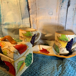 Puede incluir: Un juego de servir de cerámica con diseño de uvas y hojas. El juego incluye una caja rectangular con tapa y asa, dos tazas y una bandeja. Las tazas y la caja tienen un fondo beige moteado con ilustraciones coloridas de uvas y hojas.