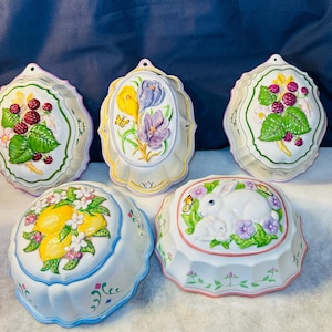 May include: Five white ceramic moulds with embossed designs. The moulds feature spring-themed motifs, including raspberries, lemons, flowers, and a bunny with butterflies. Each mould has a scalloped edge and is painted with pastel colours.