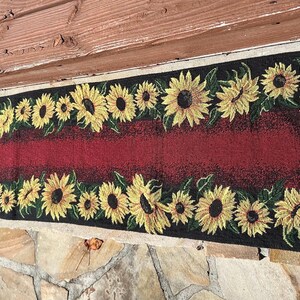 Può includere: Un runner da tavola rettangolare caratterizzato da un pannello centrale rosso scuro bordato da una cornice nera decorata con un motivo ripetitivo di girasoli gialli con foglie verdi. Il tessuto presenta una trama intrecciata ed è esposto.