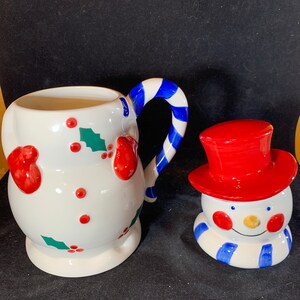 May include: A white ceramic snowman mug with a red and blue candy cane handle. The mug is decorated with holly and red berries. A separate snowman figurine with a red hat and blue scarf is also included.