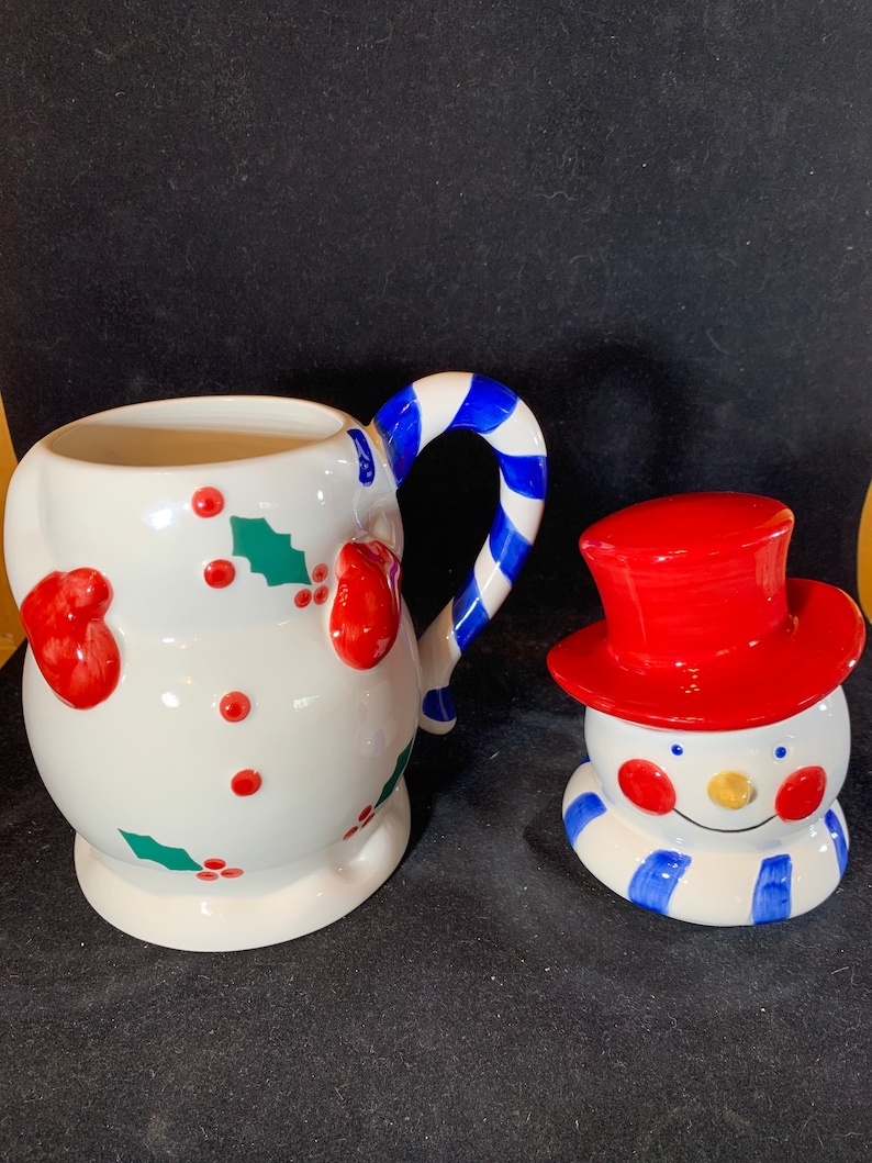 May include: A white ceramic snowman mug with a red and blue candy cane handle. The mug is decorated with holly and red berries. A separate snowman figurine with a red hat and blue scarf is also included.