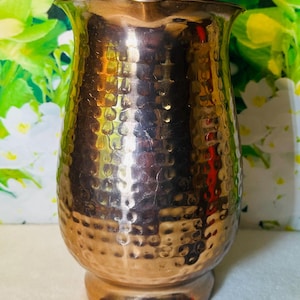 May include: A hammered copper pitcher with a wide, flared top and a rounded base. The copper has a warm, reddish-brown hue and a textured surface. The pitcher is designed with a classic, elegant shape, suitable for serving beverages.