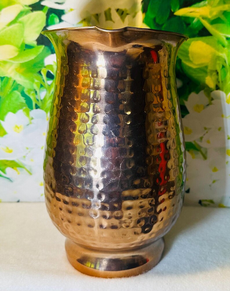 May include: A hammered copper pitcher with a wide, flared top and a rounded base. The copper has a warm, reddish-brown hue and a textured surface. The pitcher is designed with a classic, elegant shape, suitable for serving beverages.