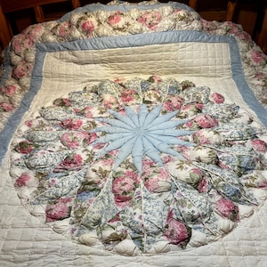 May include: A quilted bedspread featuring a central circular medallion with three-dimensional fabric petals in light blue and floral patterns of pink, white, and blue. The border is light blue, and the background is a cream-colored quilted fabric.