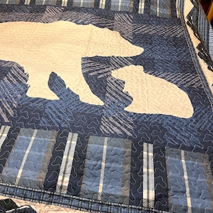 Op de afbeelding: Een blauw-witte quilt met een beer- en welp-silhouet. De quilt heeft een geruit en gestructureerd patroon in blauw, wit en grijs. De berensilhouetten zijn crèmekleurig. De quilt is geschikt voor een bed of bank.