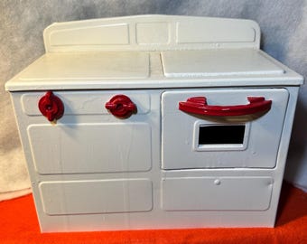 May include: A white toy stove with red accents. The stove has two red knobs, a red handle, and a black rectangular opening. The stove is sitting on a red surface.