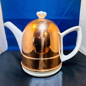 Puede incluir: Una tetera de color cobre con asa, boquilla y base blancas. La tapa es blanca con un pequeño pomo decorativo. La tetera tiene un cuerpo redondeado y una superficie pulida y reflectante. Un artículo de cocina clásico.