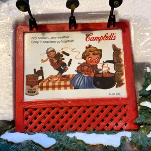 May include: A red and white Christmas village scene with a miniature Campbell's Soup sign featuring a vintage advertisement with the text "Any season...any weather...Soup 'n crackers go together!" and the Campbell's Soup logo. The sign is lit by two small black streetlights. The scene is set on a snowy base with green foliage.