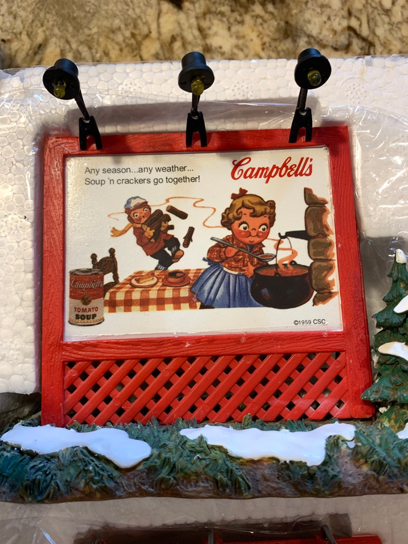 May include: A red and white Christmas village scene with a miniature Campbell's Soup sign featuring a vintage advertisement with the text "Any season...any weather...Soup 'n crackers go together!" and the Campbell's Soup logo. The sign is lit by two small black streetlights. The scene is set on a snowy base with green foliage.