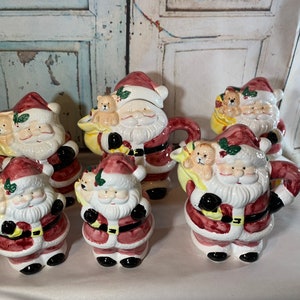 May include: Set of six ceramic Santa Claus figurines, each holding a teddy bear. The figurines are painted in red, white, and green with details of holly and a black belt.