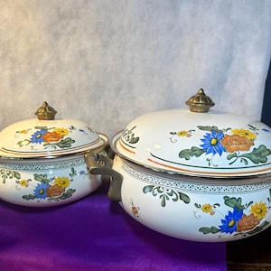 May include: Two white enamel pots with lids. Each pot features a floral design with blue, yellow, and orange flowers, green leaves, and gold trim. The pots have brass-colored handles and lid knobs, and are likely for cooking.