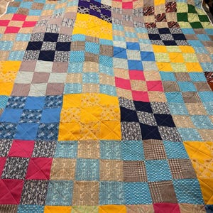 Könnte beinhalten: Eine Patchwork-Quiltdecke mit einer Vielzahl von Stoffen in einem Rastermuster. Die Quiltdecke zeigt Quadrate aus blauen, gelben, rosa und gemusterten Stoffen. Die Quiltdecke ist handgefertigt und hat ein gestepptes Design.