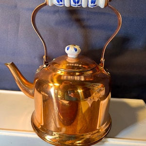 May include: A polished copper tea kettle featuring a curved spout and a rounded body. The handle is accented with a white ceramic grip decorated with blue leaf patterns, and the lid includes a matching white ceramic knob with blue floral details.