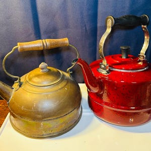 May include: Two vintage teapots. One is a tarnished brass color with a wooden handle, the other is a glossy red with a black handle. Both have spouts and lids, and are set against a blue backdrop.