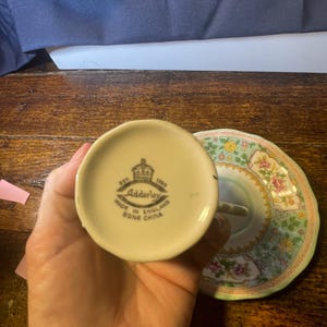 Może przedstawiać: Zestaw do herbaty z porcelany kostnej w stylu vintage z kwiatowym spodkiem. Na spodzie filiżanki widnieje napis "Adderley Made in England Bone China" i logo korony. Spodek ma zielony i r&oacute;żowy wz&oacute;r kwiatowy.